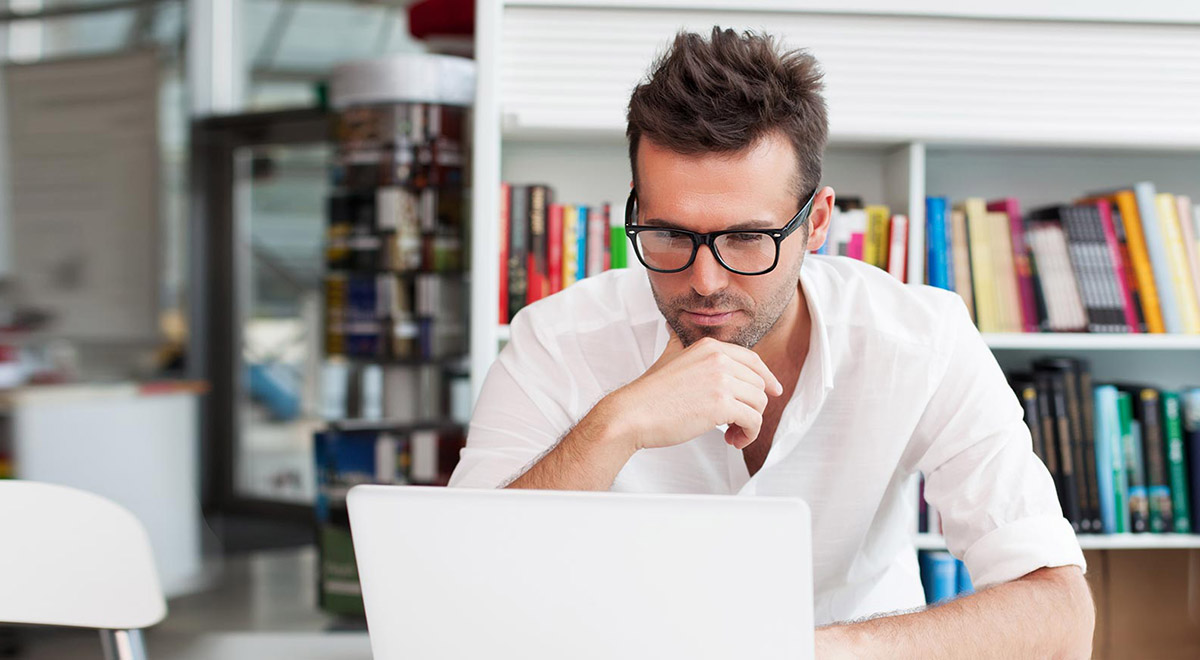 Mann mit Brille und weißem Hemd arbeitet an einem Laptop und sitzt vor einem Bücherregal.